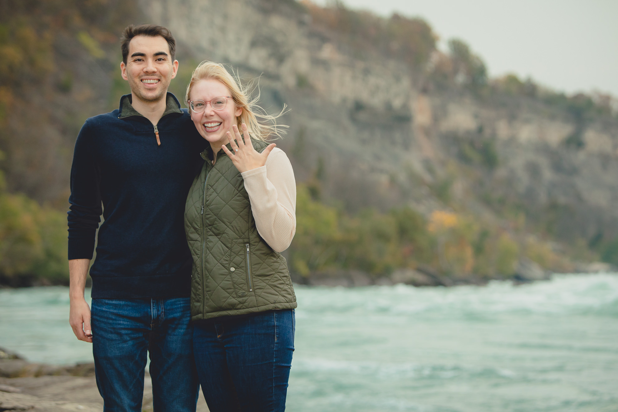 bride to be shows off engagement ring after wedding engagement proposal photography with rapids and fall foliage in background at Whirlpool State Park in Niagara Falls, NY