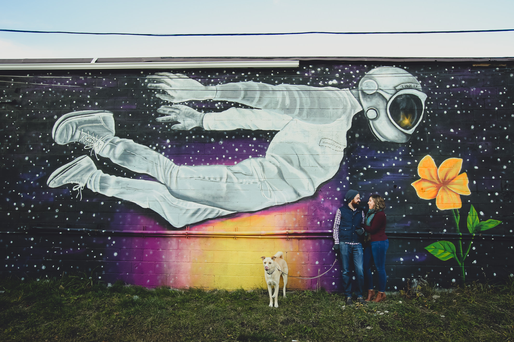 couple and dog smile at one another in front of Buffalo mural during couples mini photography session on Hertel Ave. in North Buffalo, NY