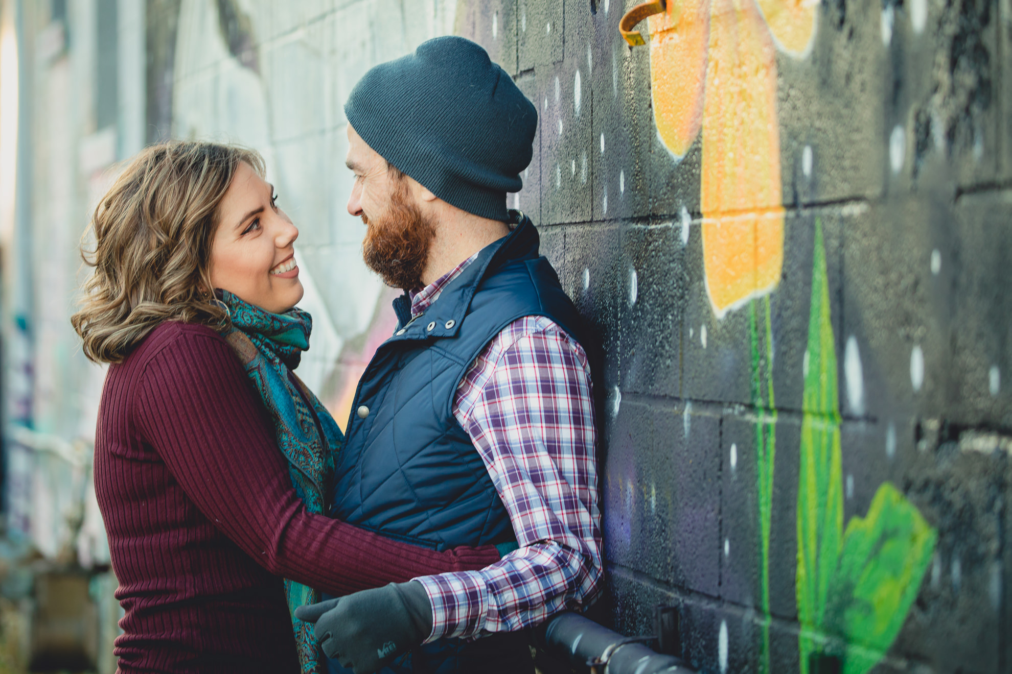 couple smiles at each other in front of Buffalo mural during couples mini photography session on Hertel Ave. in North Buffalo, NY