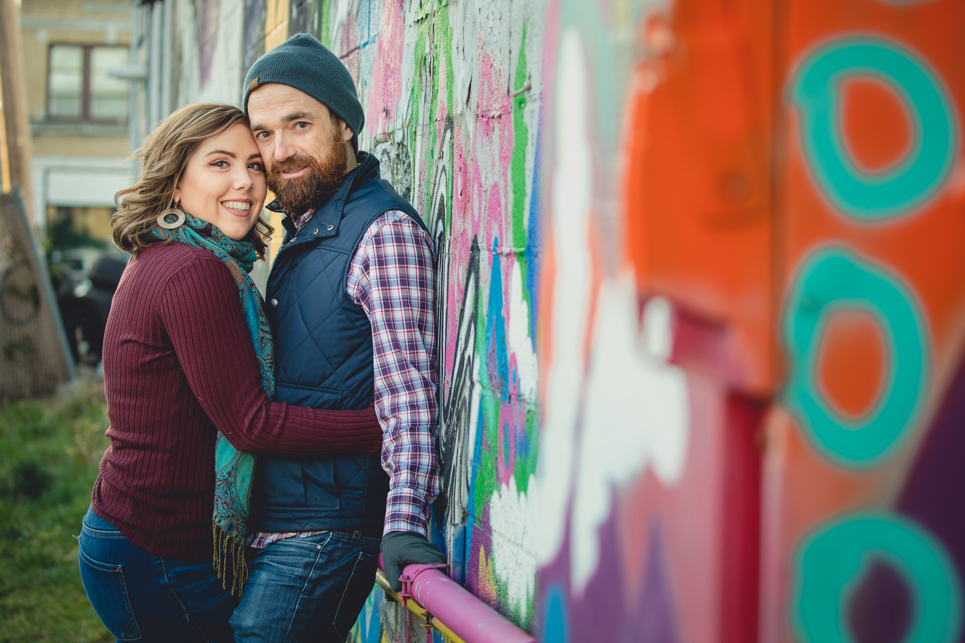 couple smiles for photographer in front of Buffalo mural during couples mini photography session on Hertel Ave. in North Buffalo, NY