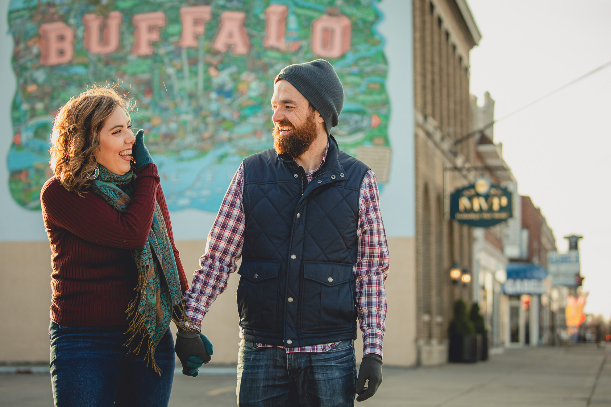 couple laughs while holding hands in front of Buffalo mural on Hertel Ave. in North Buffalo, NY during couples photography session