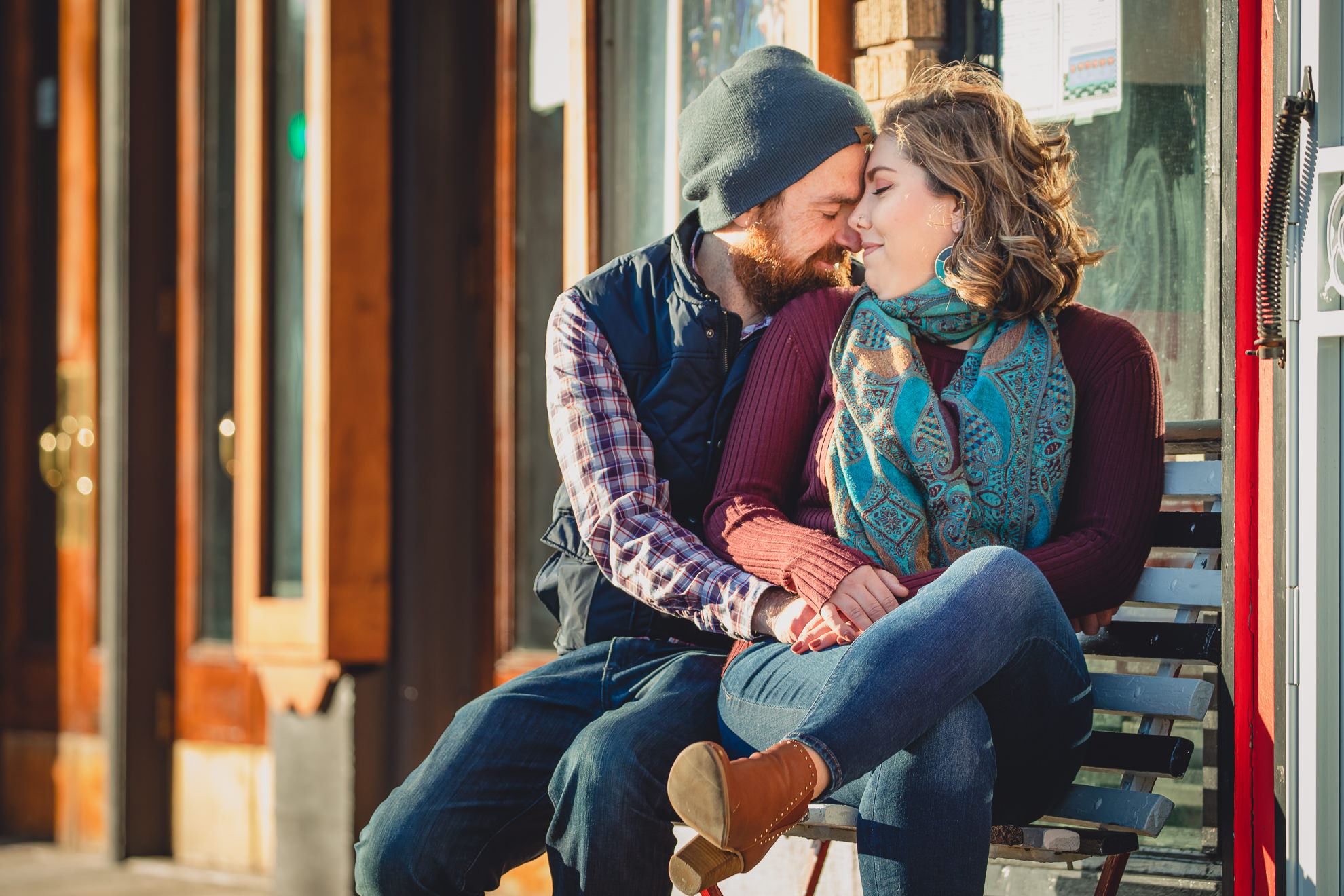 couple smiles at each other on bench in front of North Park Theater during couples mini photography session on Hertel Ave. in North Buffalo, NY