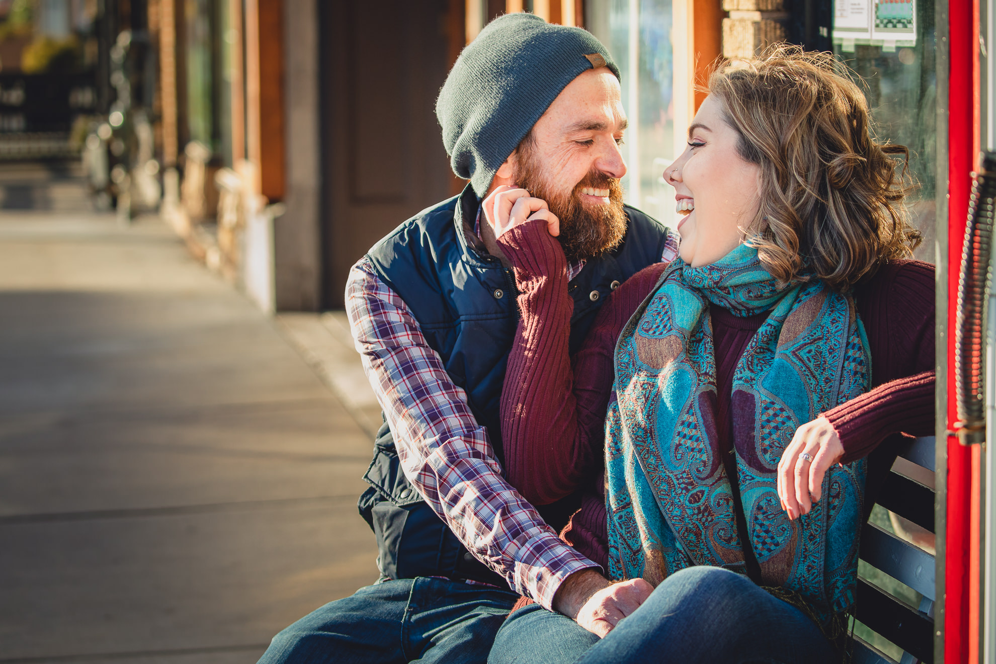 couple smiles at each other on bench in front of North Park Theater during couples mini photography session on Hertel Ave. in North Buffalo, NY