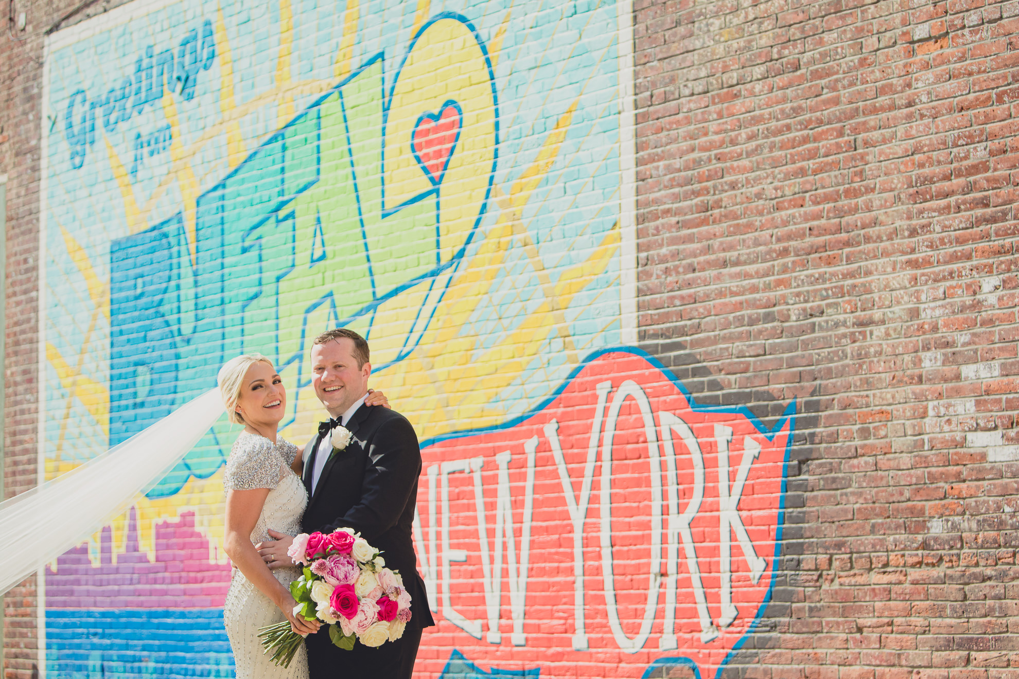 Bride and groom pose for wedding portrait in front of Greetings from Buffalo mural the theater district of down town Buffalo.