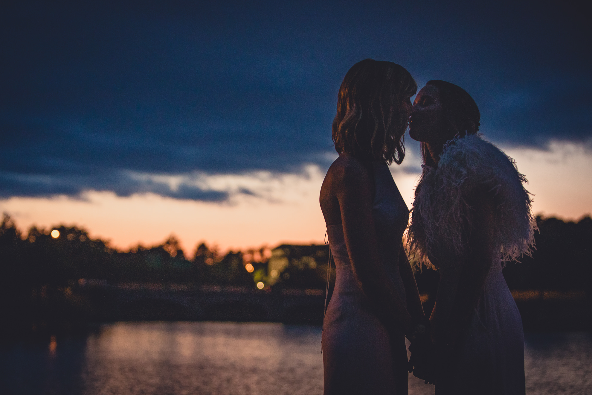 brides kiss in front of sunset over Hoyt Lake during wedding at Marcy Casino in Delaware Park in Buffalo, NY