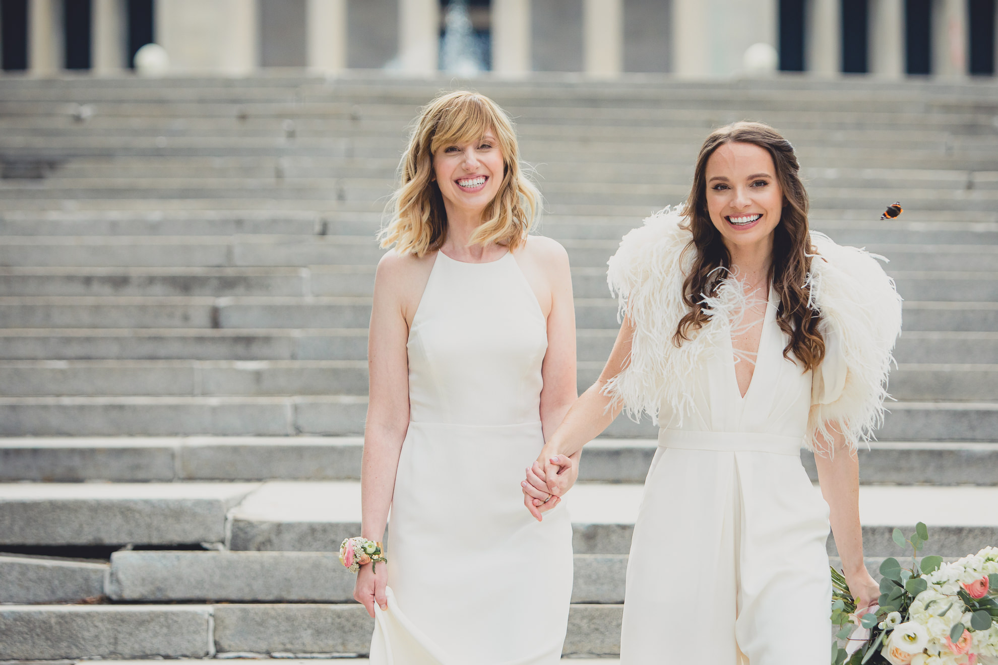 brides laugh as butterfly swarms on steps to Hoyt Lake during portrait session on wedding day in front of Albright Knox in Delaware Park in Buffalo, NY