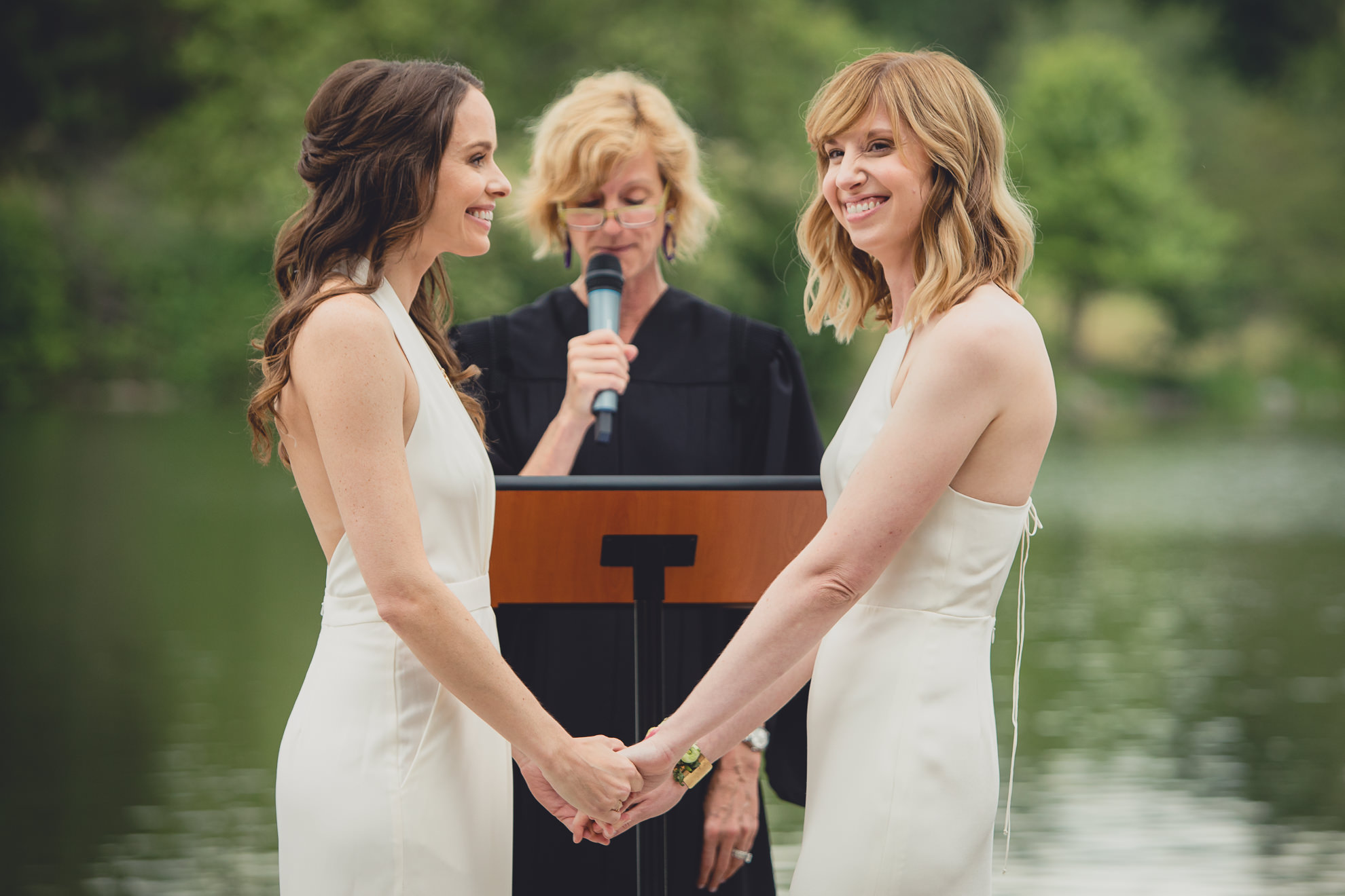 brides smile at guests during wedding ceremony in front of Hoyt Lake at Marcy Casino in Delaware Park in Buffalo, NY
