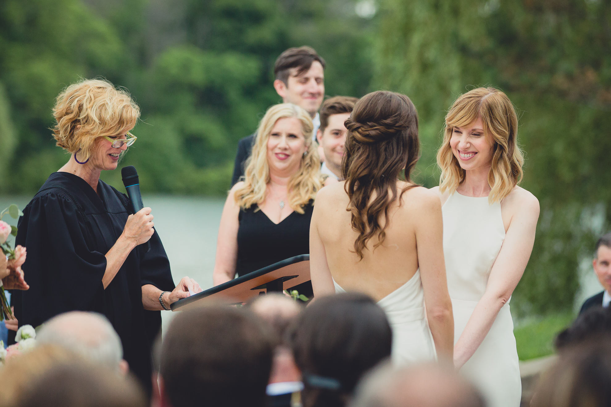 brides smile at each other during wedding ceremony in front of Hoyt Lake at Marcy Casino in Delaware Park in Buffalo, NY