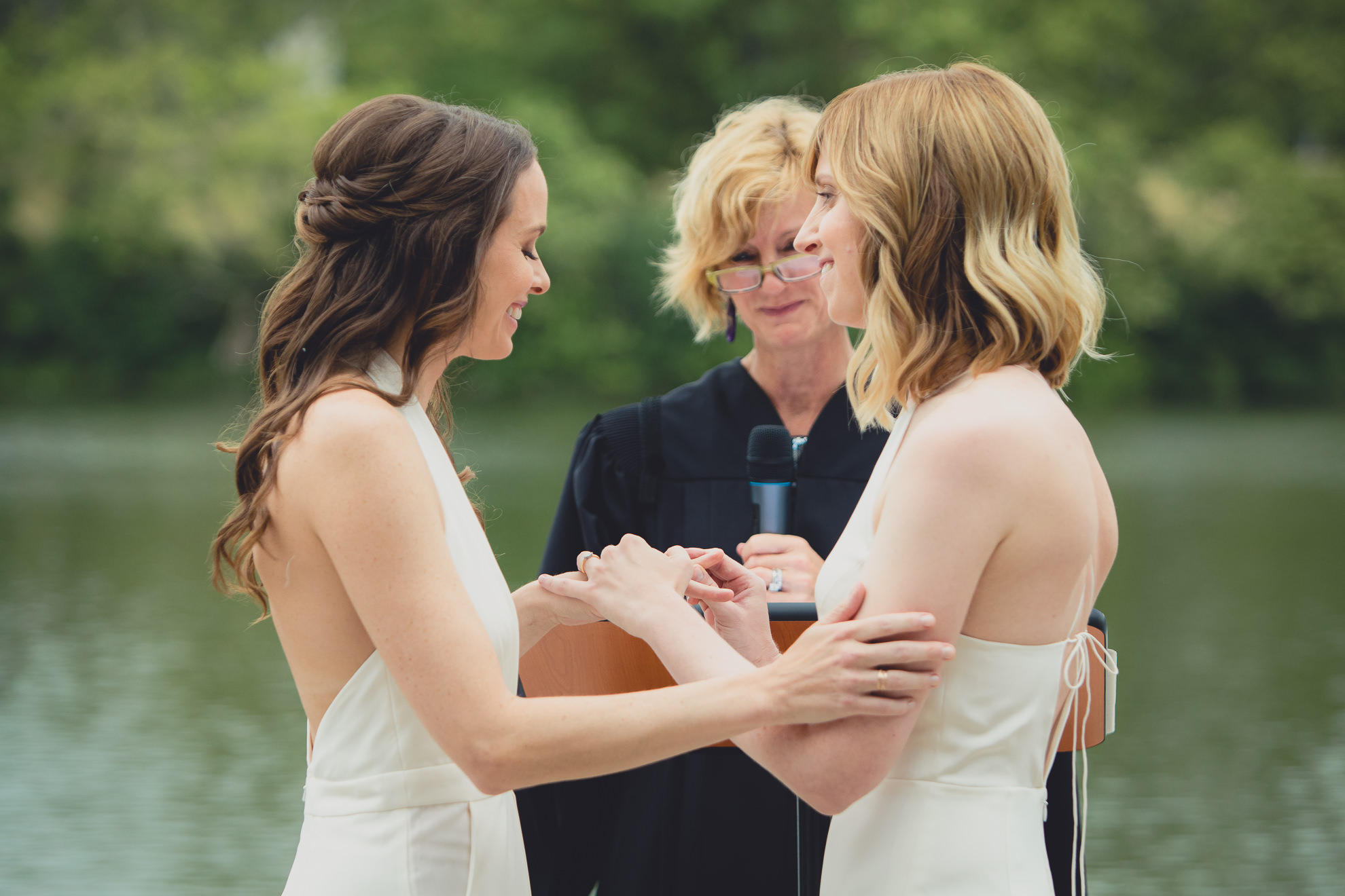 brides place rings on each others fingers during wedding ceremony in front of Hoyt Lake at Marcy Casino in Delaware Park in Buffalo, NY