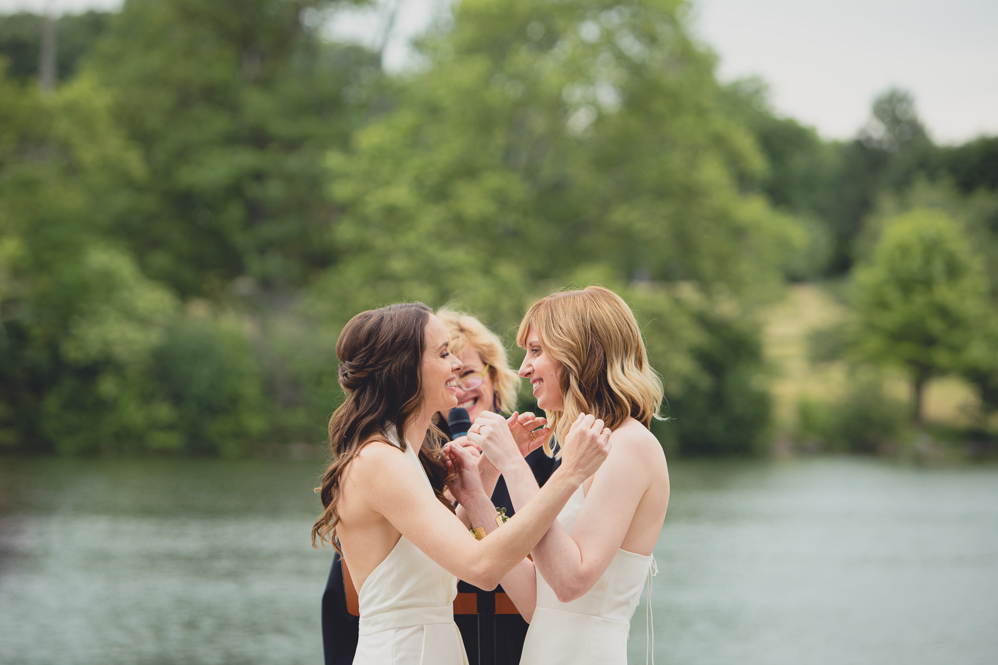 brides smile at one another during wedding ceremony in front of Hoyt Lake at Marcy Casino in Delaware Park in Buffalo, NY
