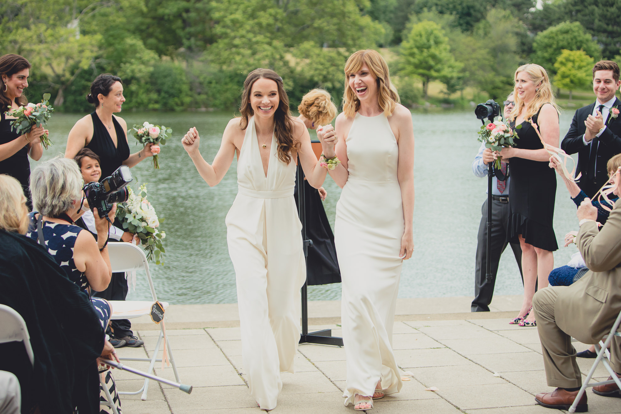 brides celebrate during recessional during wedding ceremony in front of Hoyt Lake at Marcy Casino in Delaware Park in Buffalo, NY