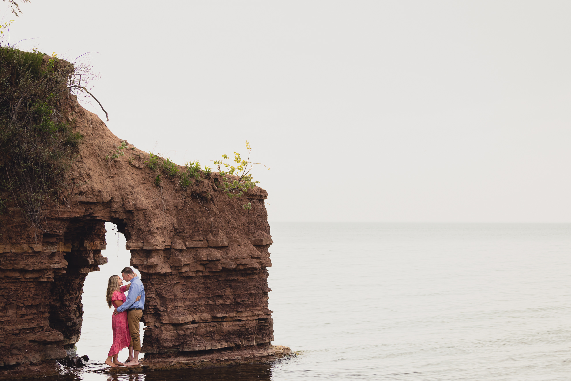 couple looks at each other under archway in cliff on Lake Ontario beach during wedding engagement proposal photography in Niagara County