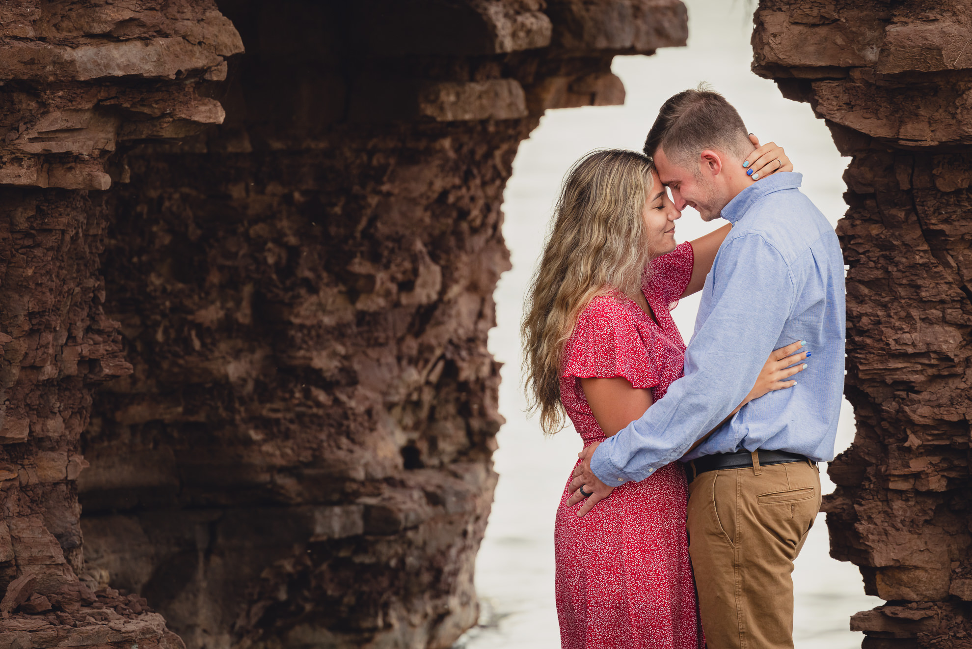 couple looks at each other under archway in cliff on Lake Ontario beach during wedding engagement proposal photography in Niagara County