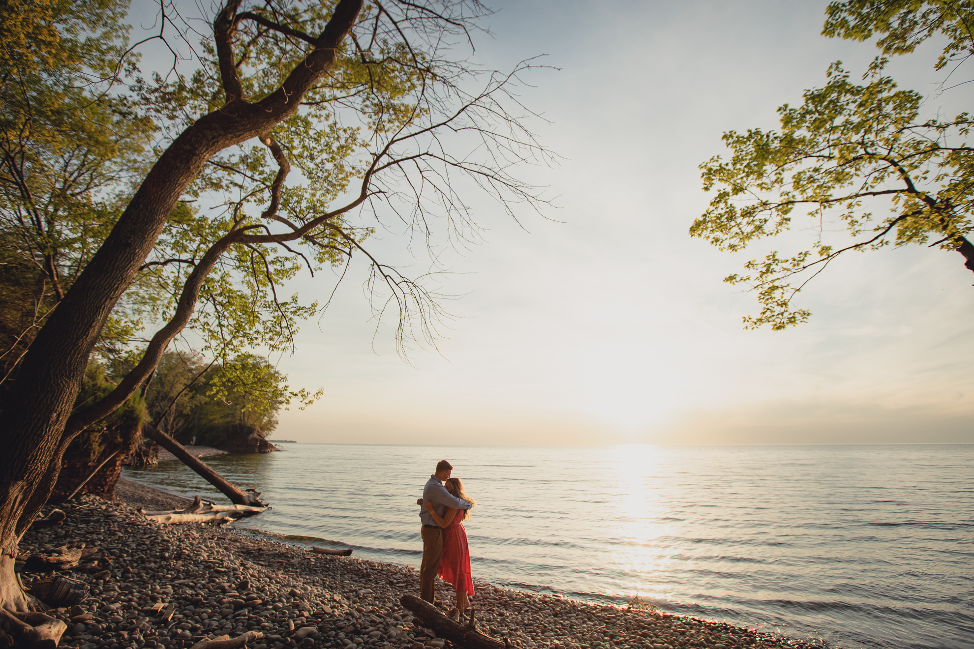 couple snuggles during proposal on Lake Ontario beach at sunset during wedding engagement proposal photography in Niagara County