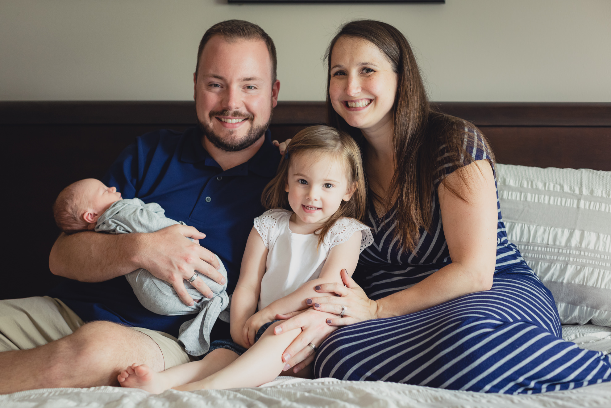 family smiles for photographer while sitting on bed during newborn baby family photography session in Buffalo, NY