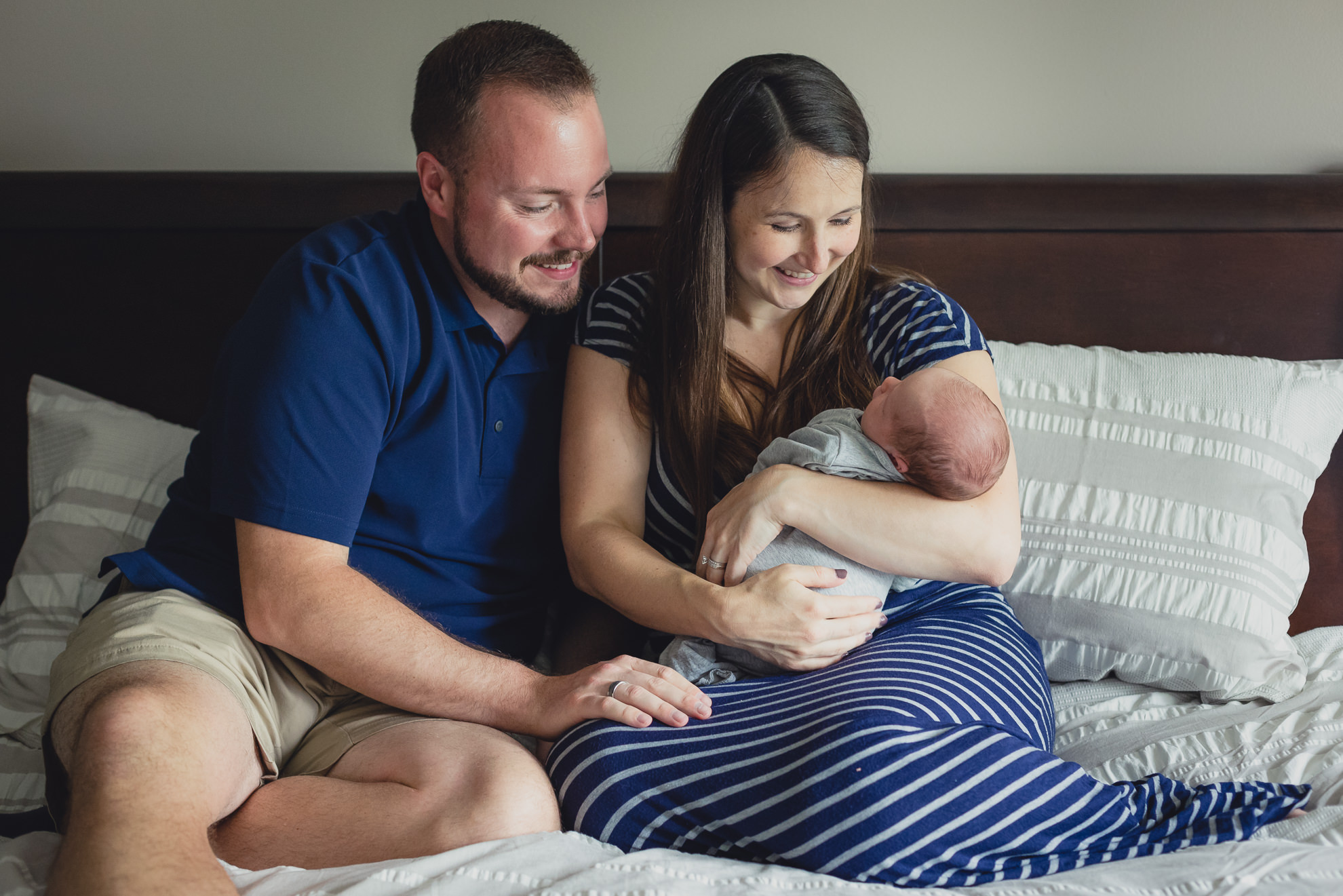mom and dad look at baby while sitting on bed during newborn baby family photography session in Buffalo, NY