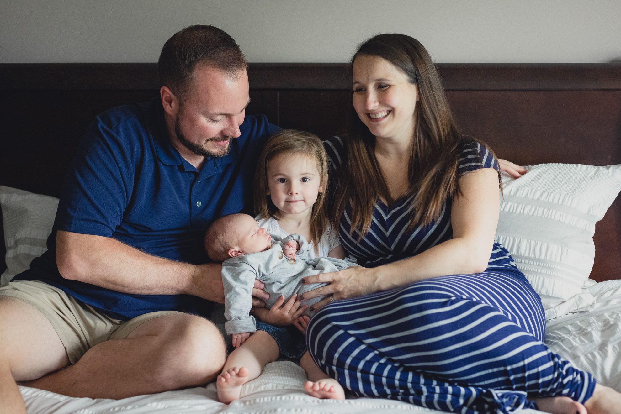 family looks at each other while sitting on bed during newborn baby family photography session in Buffalo, NY