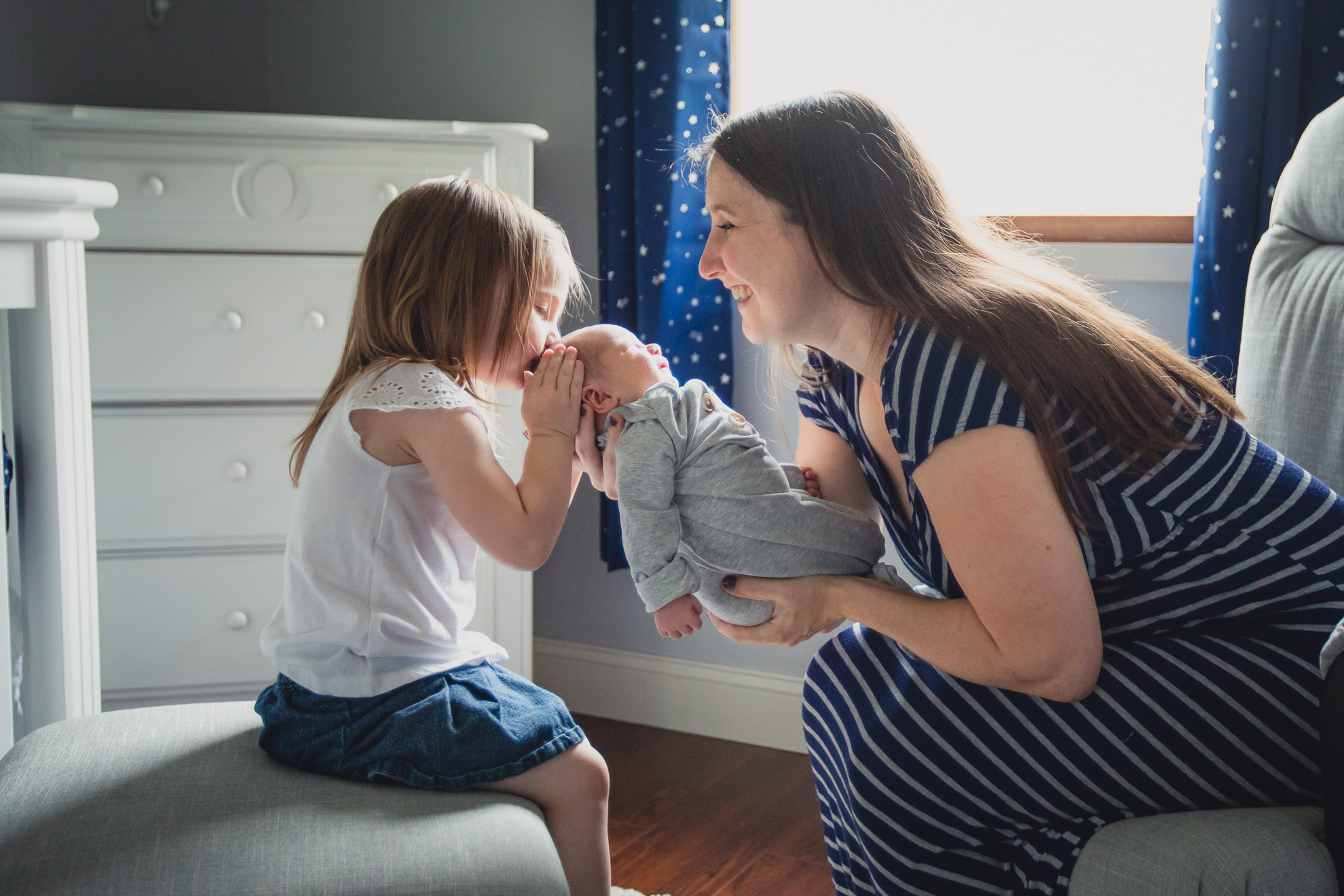 big sister kisses baby mom is holding in nursery while sitting on bed during newborn baby family photography session in Buffalo, NY
