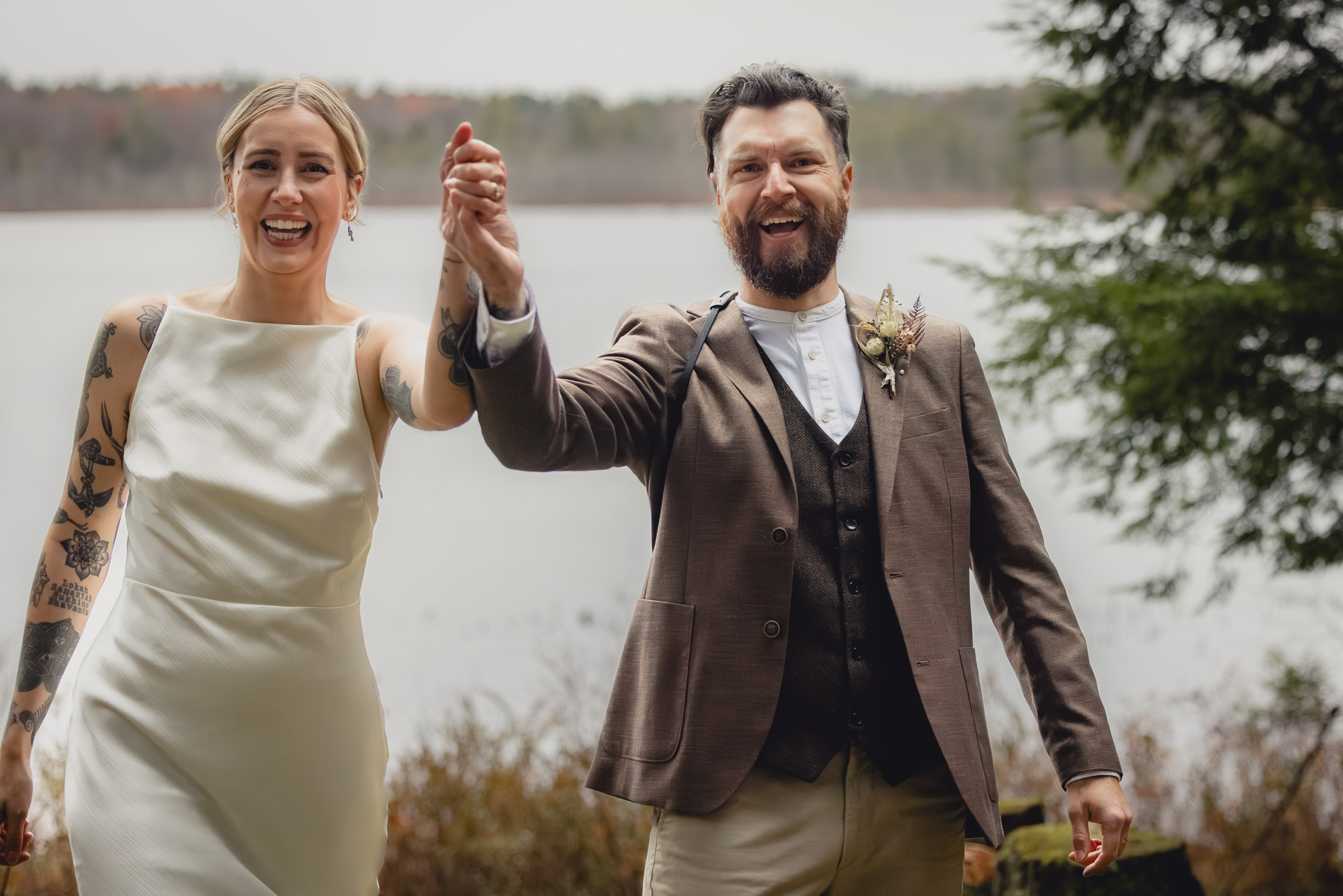 bride and groom raise arms in celebration in front of lake during micro wedding elopement ceremony at Vanderkamp NY in the Adirondacks