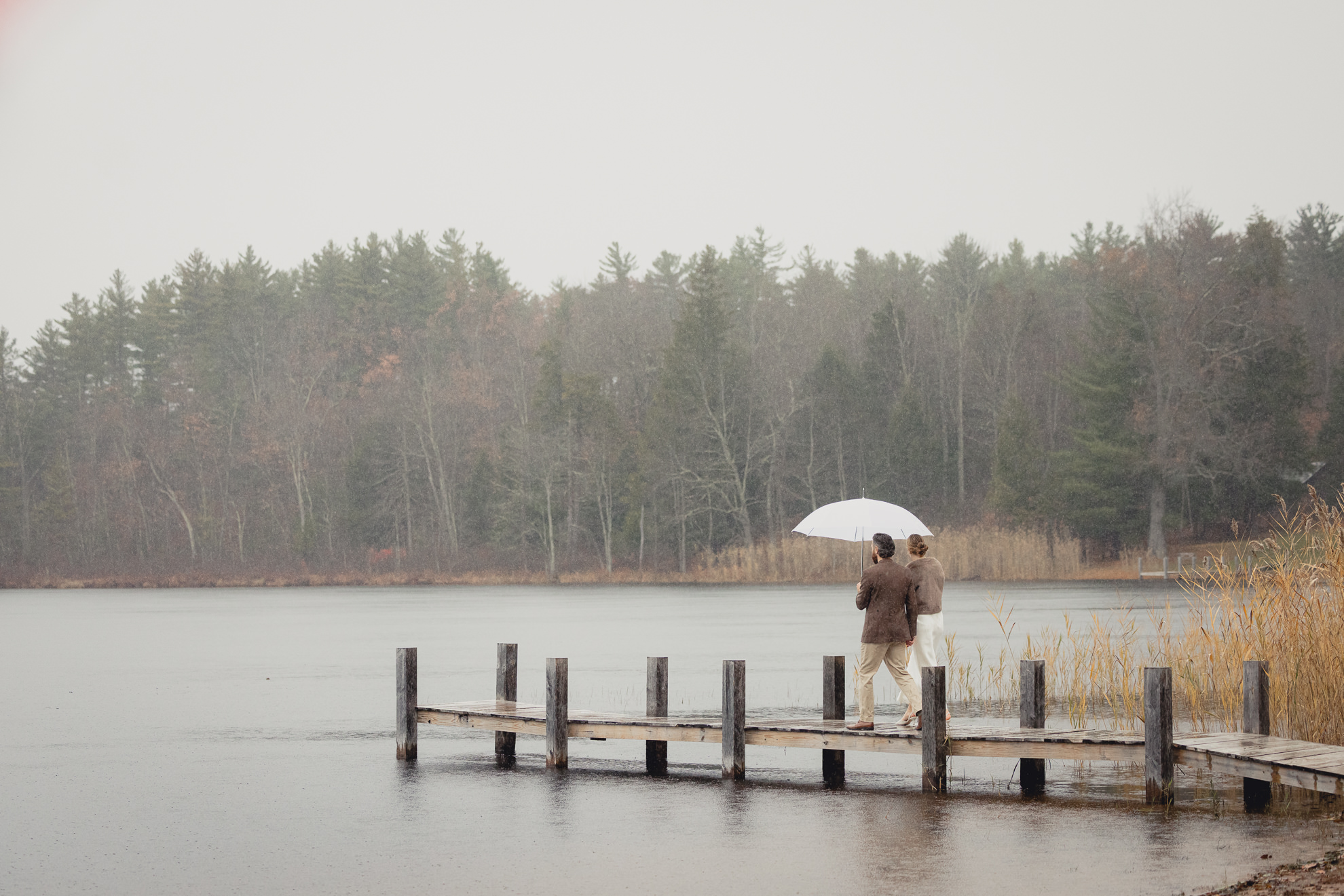 bride and groom walk down dock at lake with umbrella during fall after micro wedding elopement ceremony at Vanderkamp NY in the Adirondacks