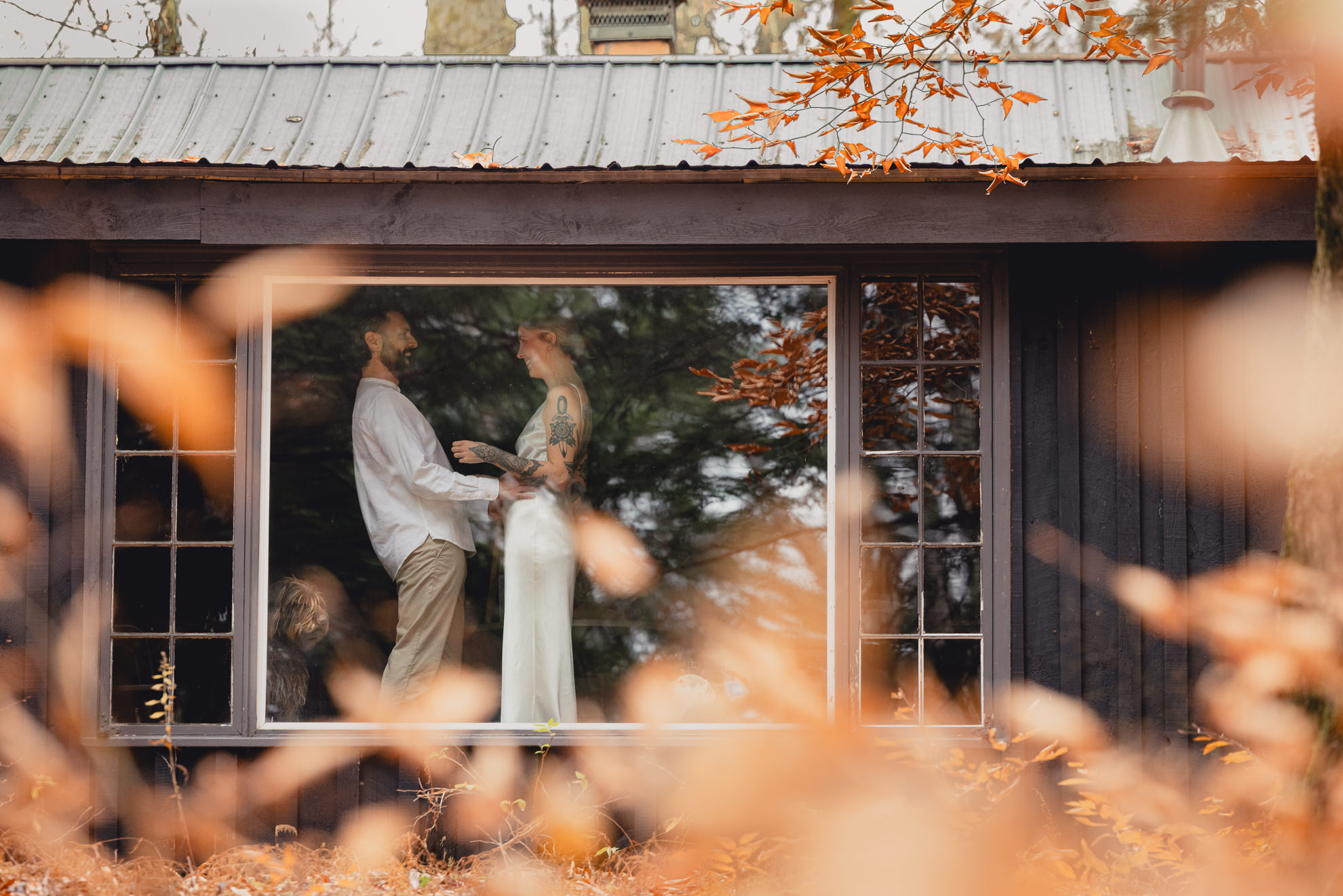 groom is seen helping bride get ready through window in Langston Cottage at Vanderkamp NY before fall wedding elopement in Adirondacks